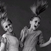 Two women in light-colored dresses with high ponytails pose against a dark background, one waving with a raised hand.