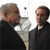 Two men in dark suits stand side-by-side against a bright background, one with gray hair and the other with brown hair, both facing forward.