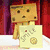 A pixelated Danbo character holds a yellow card with a flower and coin design against a red curtain background.