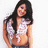 A woman with long dark hair wears a white patterned crop top and blue jeans, smiling against a plain background.