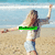 A pixelated beach scene shows a person with long blonde hair in a white top and light blue shorts, arms outstretched on sand with ocean and sky background.