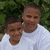 Two people in white shirts pose closely together against a green leafy background, with one person slightly behind the other.