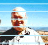 A person in a light-colored shirt with dark accents stands against a bright blue sky and distant landscape, partially obscured by horizontal light blue bars.