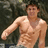 Pixelated image of a shirtless man with light brown hair standing against a dark background, his right arm extended forward.