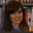 A person with long brown hair and a blue top smiles in front of bookshelves filled with books.