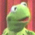 Green frog-like puppet with large eyes and a wide mouth against a red background, rendered in pixelated style.