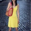 A person in a yellow sleeveless dress holding a red and beige handbag with a patterned design stands on a cobblestone street.