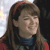 A person with long brown hair and a red headband wears a dark blue top with red accents, set against a light background with a dark area on the right.