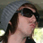 A person wearing a gray knit beanie and dark sunglasses, with long brown hair tied back in a braid, shown from the shoulders up against a blurred outdoor background.