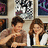 Two people sit at a table in front of framed artwork, with text reading Music And Lyrics and <333333 on blue