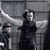 Two pixelated figures in dark clothing stand against a gray grid background; one figure extends an arm toward the other.