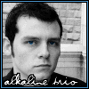 A black-and-white portrait of a person with short dark hair and blue eyes, wearing a dark shirt, with 'alkaline trio' text below.