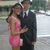 A static image of a couple in formal attire standing outdoors, the woman in a pink dress and the man in a black suit with a white shirt and black tie.