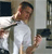 A person in a white shirt holds a dark rectangular object near their face with one hand while extending the other arm outward, set against a blurred background with dark and light areas.