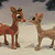 Two reindeer stand side-by-side on a snowy ground with a blurred background, one larger and one smaller.