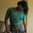Pixelated image of a person with dark hair wearing a teal shirt and dark pants, standing against a light brown background.