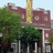 Pixelated static image of a building with red brick facade, multiple windows, and a tall yellow chimney, partially obscured by green foliage.