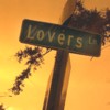 A street sign reading 'Lovers' against an orange sky with blurred tree branches.