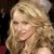 Pixelated close-up of a person with long wavy blonde hair, light skin, and a slight smile, set against a dark background with indistinct shapes.