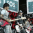Pixelated image of a band performing with red text overlay listing band name, song, and date.