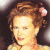 Pixelated close-up of a person with light brown hair styled in an updo featuring a pink flower accessory, wearing a light-colored top against a dark background.