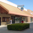 Static pixelated image of a single-story building with a light brown roof, beige walls, and dark windows under a clear blue sky.