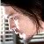 Pixelated image of a person with light skin tone holding a white object near their face, standing beside a building with dark windows.