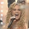 A woman with curly blonde hair sings into a black microphone, wearing an orange dress, with vertical white text reading WINNER on the left side.