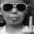 Pixelated grayscale image of a person wearing dark sunglasses and a light-colored shirt, with visible hair on the right side.
