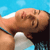 A pixelated image shows a person with light skin and brown hair leaning forward, wearing a white top with dark straps, against a bright blue background.