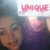 A pixelated image of a person with light blue hair wearing a purple top, holding a pink 'UNIQUE' sign, with visible text and color details.
