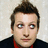 Pixelated static image of a person with short dark hair styled upward, light skin, blue eyes, and a neutral expression wearing a dark jacket over a red shirt against a light background.