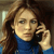 A pixelated image of a person with long brown hair wearing a blue top, holding a black phone to their ear against a blurred background.