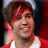 A person with short brown hair and red highlights wears a red jacket over a white shirt, smiling against a red and dark background.