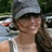 A pixelated image of a person with long brown hair wearing a gray cap and white tank top, set against a bright pink background with text elements.