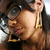 A woman with dark hair and large round glasses wears gold hoop earrings and a dark top, her head tilted slightly with a subtle smile.
