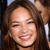 A pixelated close-up of a person with brown hair smiling broadly, showing teeth, against a dark background with a light blue element at the top left.