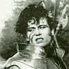 Black-and-white image of a person with curly hair wearing a high-collared garment, set against a textured background with abstract shapes.