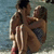 Two figures sit closely together on a beach, one figure's arm around the other in an embrace.