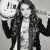 A pixelated grayscale image of a person with long hair wearing a dark jacket over a light shirt, holding up two hands in front of their face.