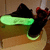 A pair of black sneakers with red accents and green glowing soles rests on a light brown surface, viewed from above.