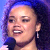 Pixelated static image of a person singing into a microphone with dark curly hair and blue lighting effects.