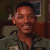 Pixelated static image of a person with closed eyes, wearing a dark green military-style uniform with visible collar and shoulder details, set against a dark background with a framed object on the right.