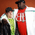 Two figures stand side-by-side against a brown background; the taller figure wears red with white text and a black cap, while the shorter figure wears green and black clothing.