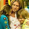 Three people embrace in a close-up; the central figure wears a white top with red text and a necklace, flanked by two others in blue and yellow patterned clothing.