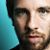 Pixelated close-up of a person's face with dark hair, light skin tone, and visible beard stubble against a teal background.