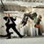 Pixelated static image shows two figures in motion against a blurred background; one figure in dark clothing appears to be kicking or striking while the other stands nearby.