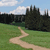 Pixelated landscape with winding dirt path through green field, dark trees, and blue sky with white clouds.