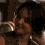 A woman with dark hair holds a microphone, smiling in a dimly lit setting with warm lighting and blurred background elements.
