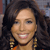 A pixelated close-up of a person with dark curly hair, wearing large gold earrings and an orange garment, smiling with visible teeth against a blurred background.